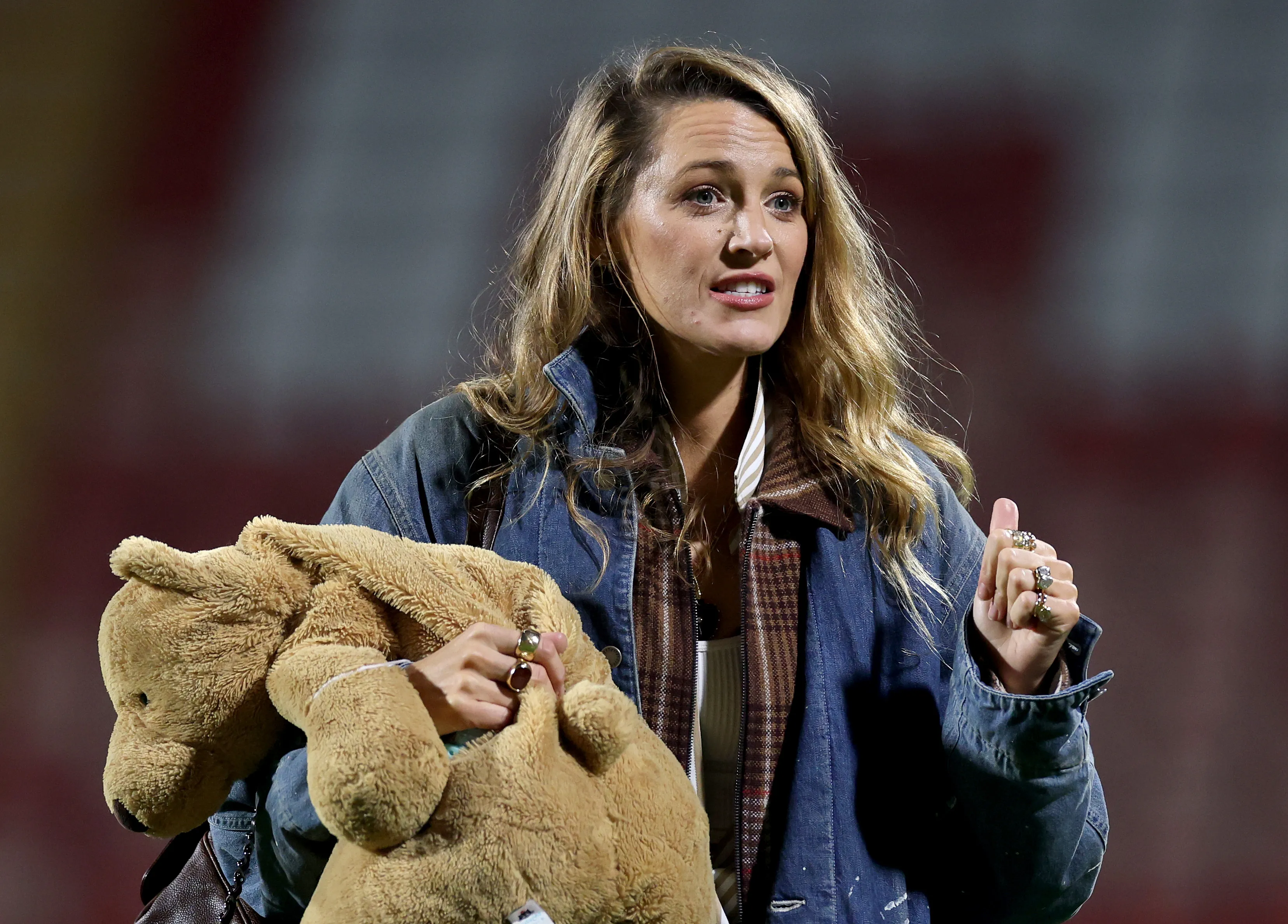 Actress Blake Lively holds a stuffed teddy bear, wearing a denim jacket over a brown plaid shirt.