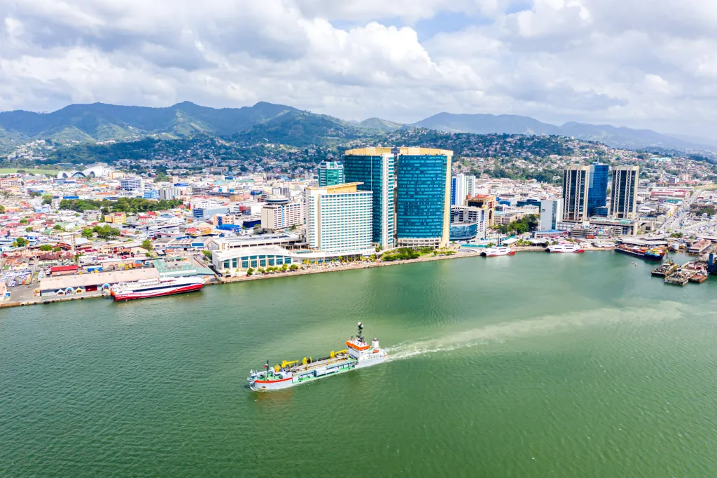 Aerial view of Port of Spain, Trinidad and Tobago, showing downtown skyscrapers, a busy seaport, and a boat leaving a wake in the water.