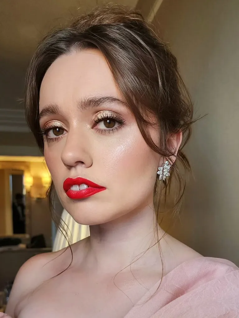 Aimee Lou Wood with a sparkly gold eyeshadow, red lipstick, and a diamond earring.