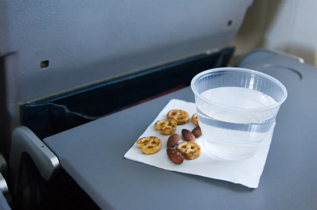 An airplane tray table with a cup of water, pretzels, and almonds on a napkin.