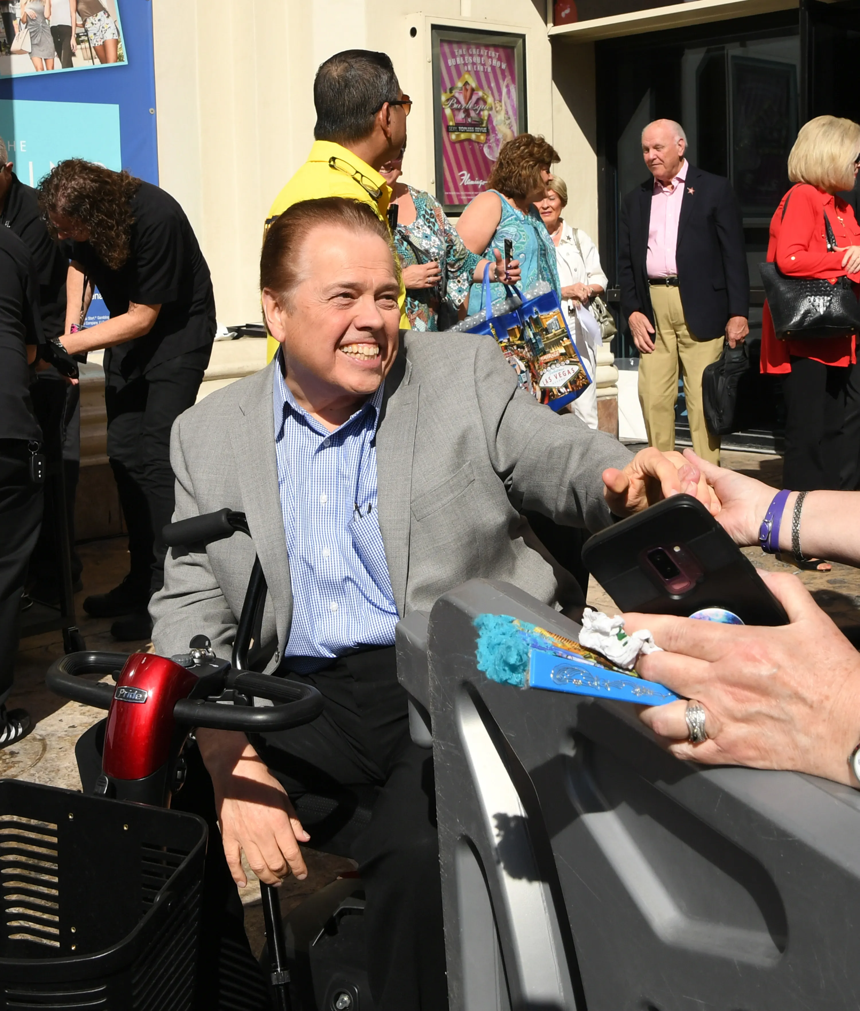 Alan Osmond seated in a motorized wheelchair, smiling while reaching out to a hand holding a smartphone.