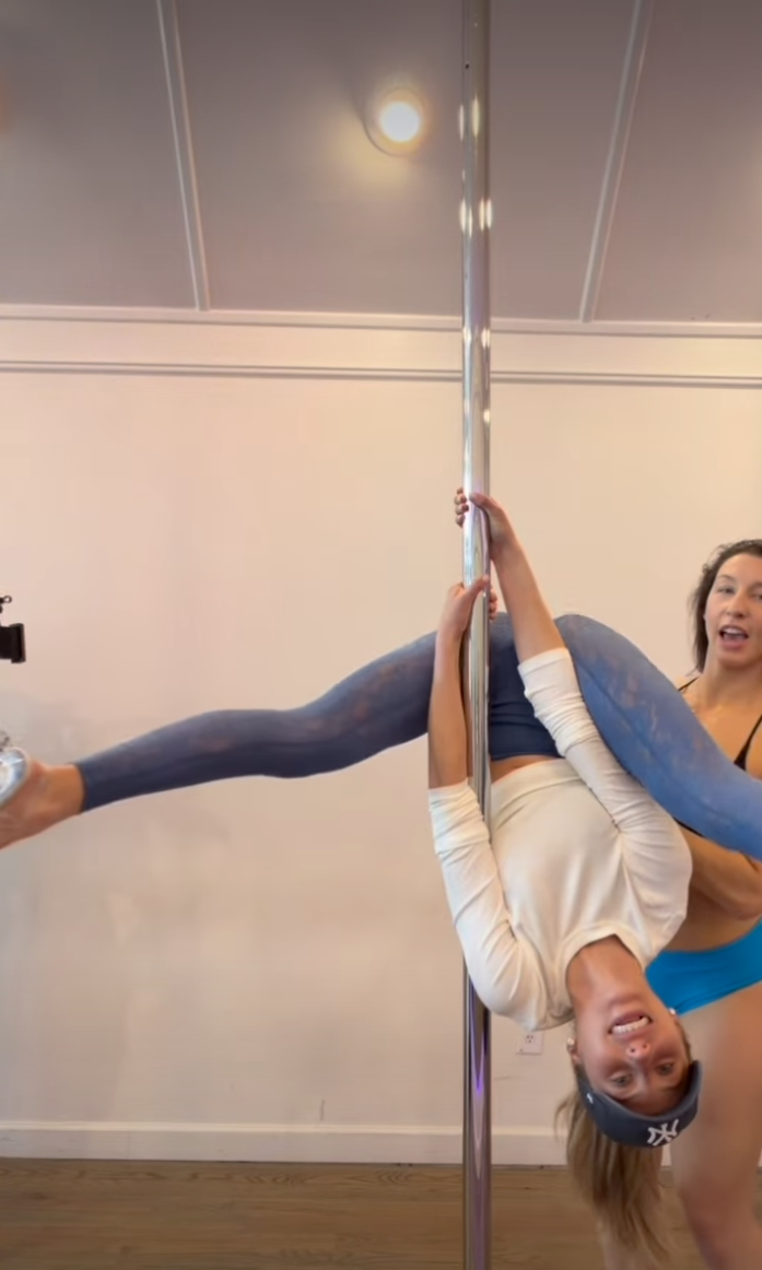 Alix Earle hanging upside down from a pole with her legs extended to the side, while a second person stands behind her.