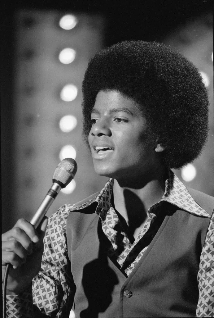 Michael Jackson with an afro and a patterned shirt, holding a microphone, performs on stage.
