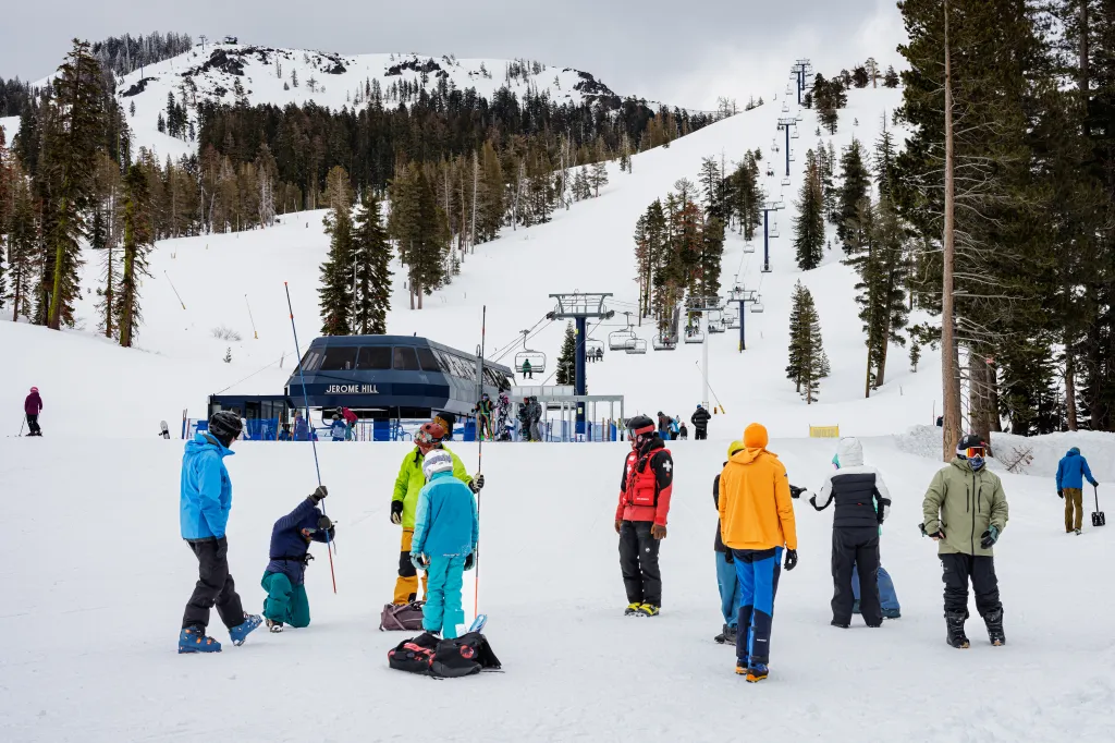 People in ski gear practice using avalanche transceivers and shovels on a snowy mountain with ski lifts.