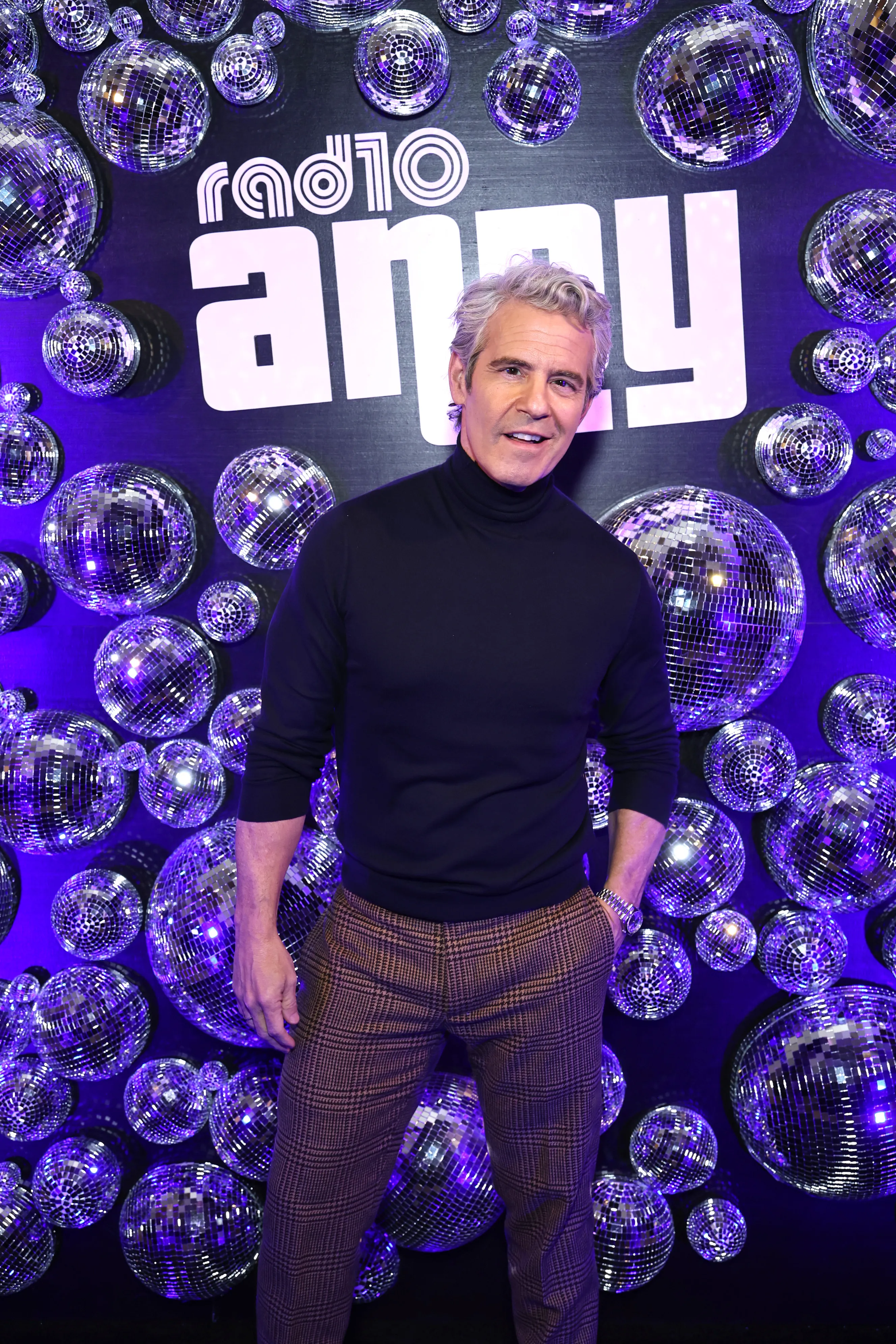 Andy Cohen in a black turtleneck and plaid pants, posing in front of a purple wall decorated with disco balls and the 