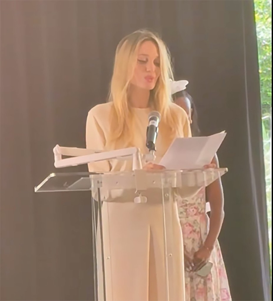 A blonde woman in an off-white outfit speaking at a podium with a microphone, while Zahara Jolie-Pitt stands next to her.