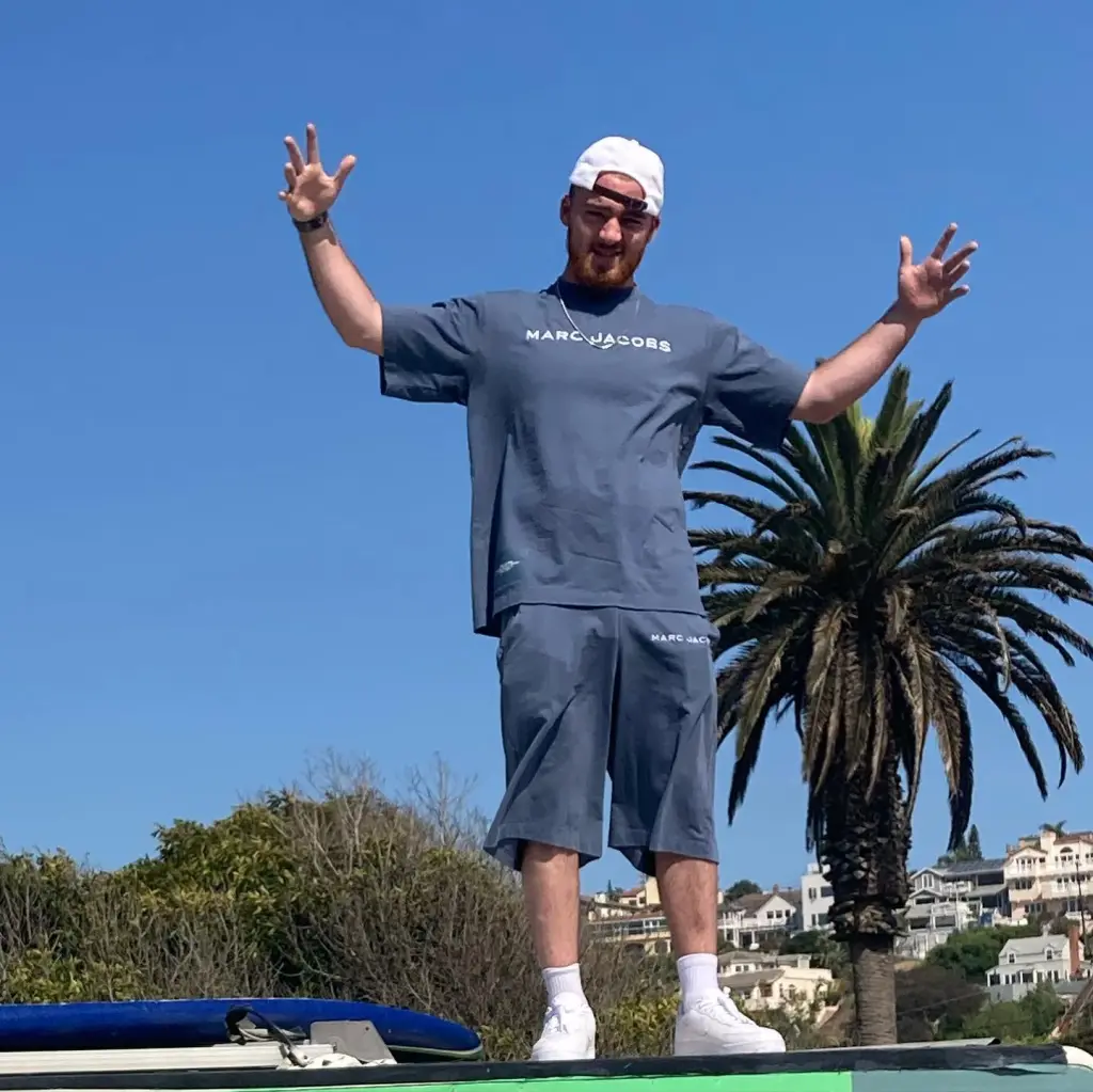 Angus Cloud wearing a gray Marc Jacobs outfit and a white cap, standing with arms raised under a clear blue sky.