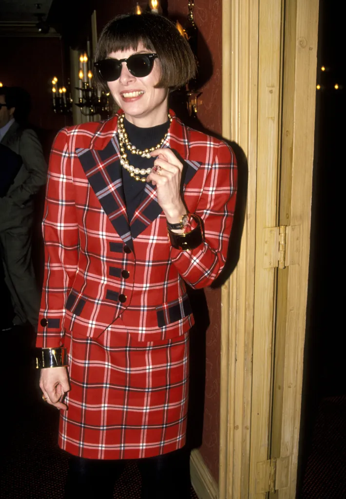Anna Wintour smiling while wearing a red plaid suit, sunglasses, and multiple necklaces.