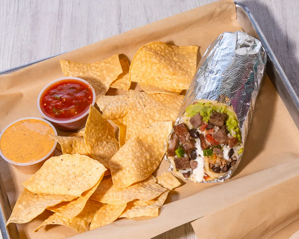 Burrito wrapped in foil with a visible cross-section of meat, rice, and beans, alongside tortilla chips and two dips.