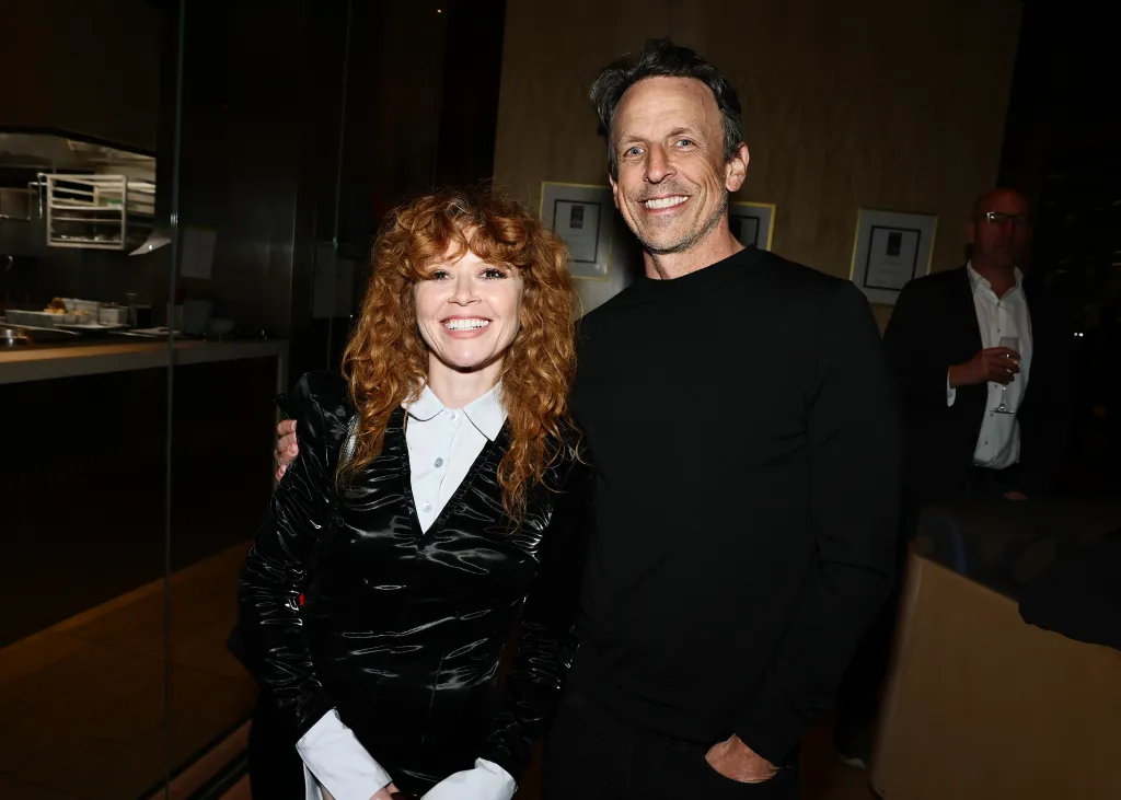 Natasha Lyonne, in a black coat and white shirt, and Seth Meyers, in a black sweater, smiling at the