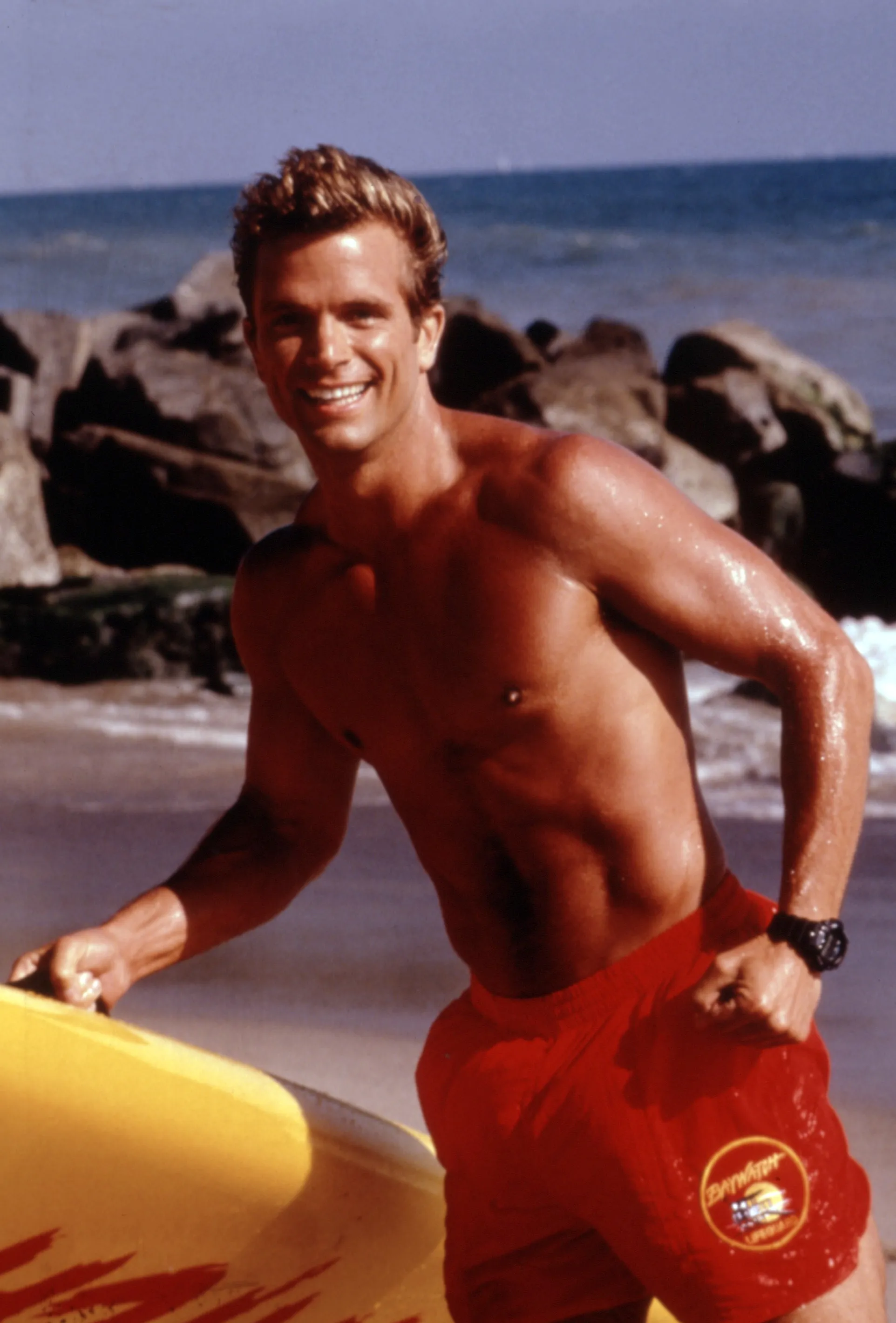 David Chokachi as a lifeguard in red shorts, smiling with a surfboard on a beach.