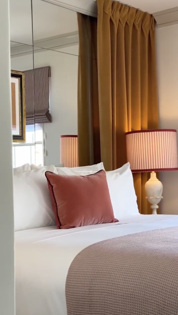 A neatly made bed in a hotel room with white sheets, a coral-pink throw pillow, and a waffle-textured blanket.