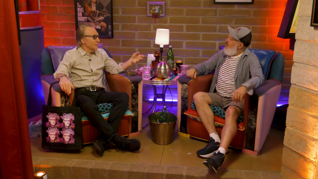 Bill Maher and David Cross sitting in chairs across from each other, engaged in conversation.