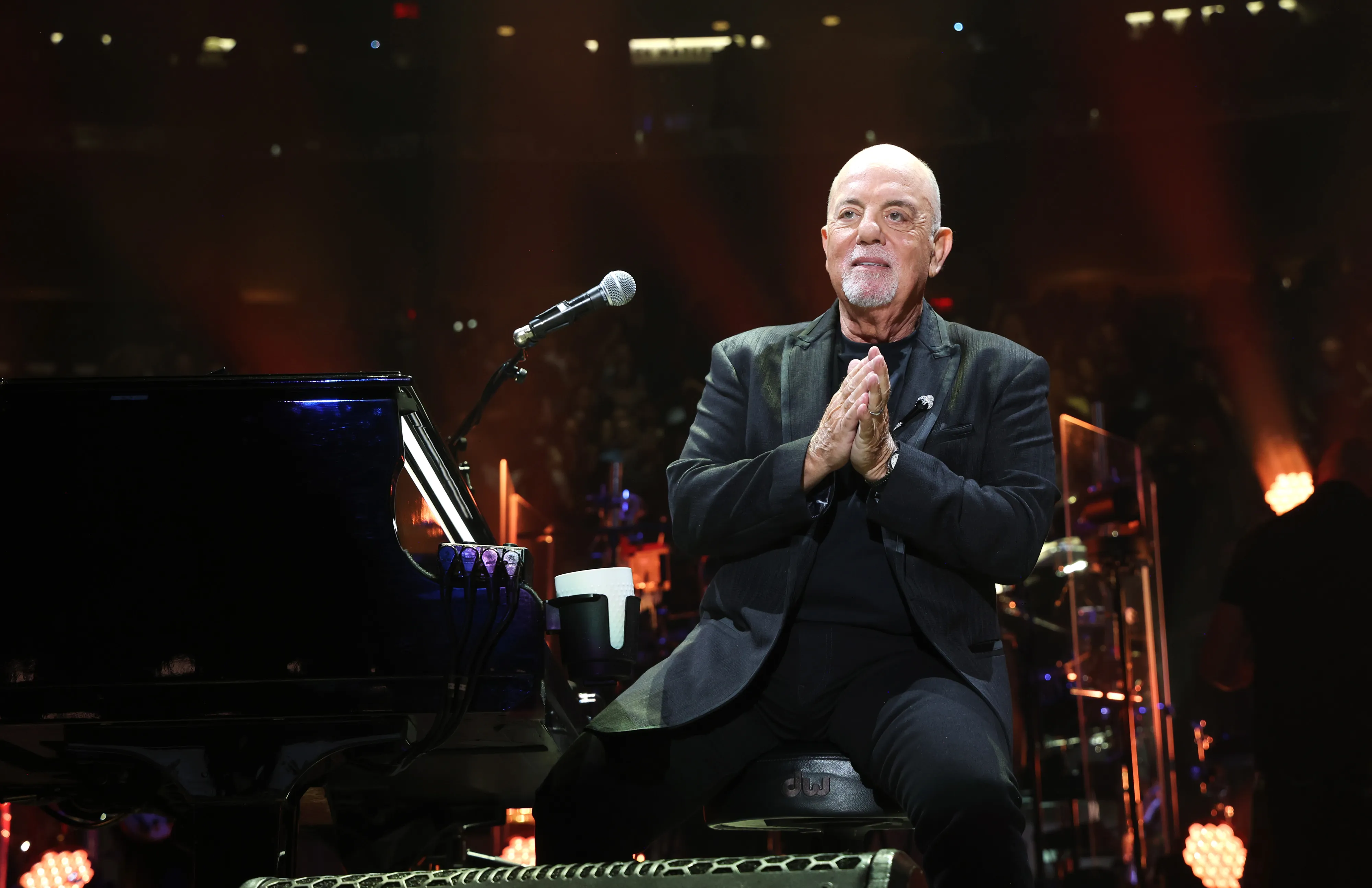Billy Joel performing on stage, sitting at a piano with his hands clasped, wearing a dark suit.