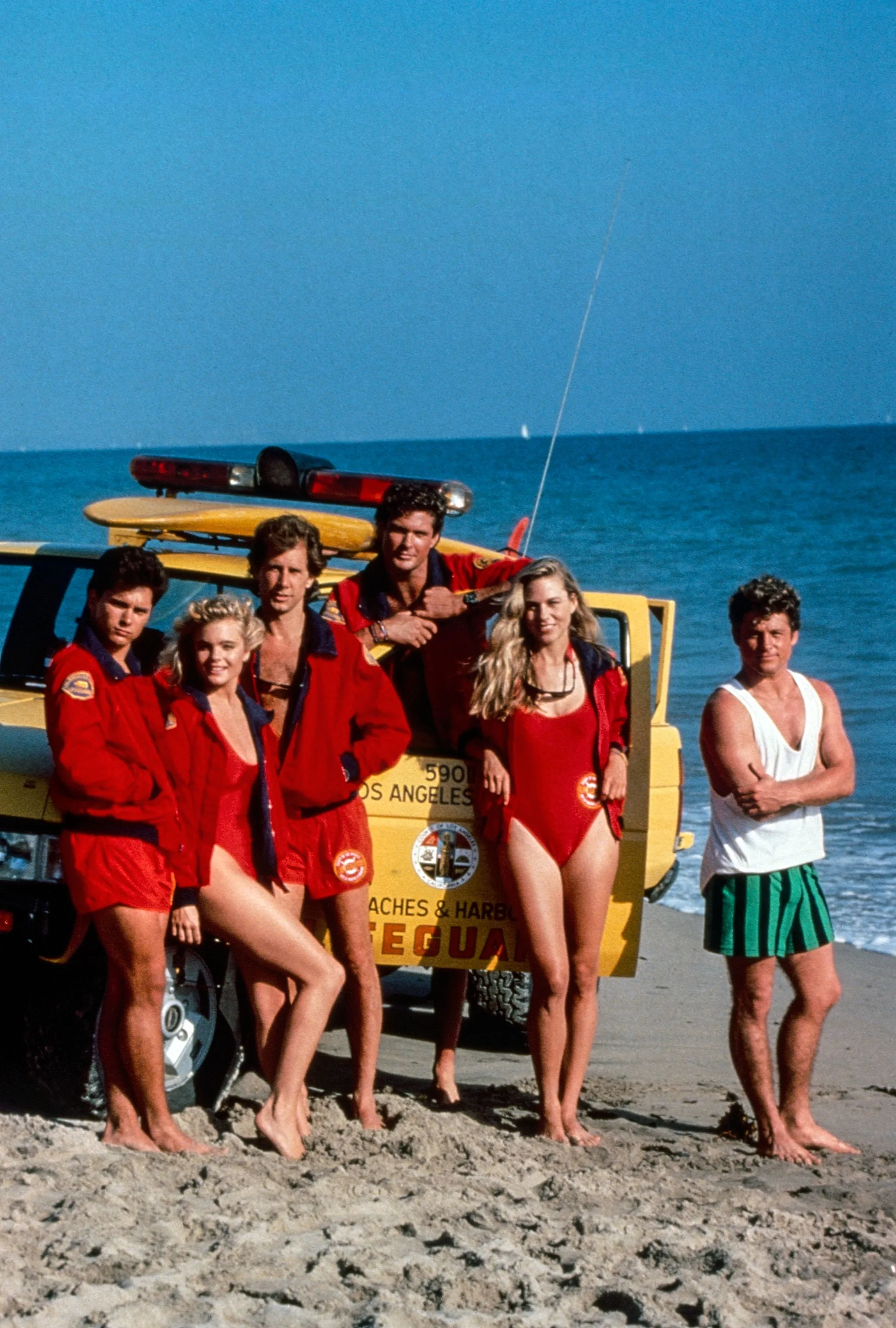 The cast of the TV show Baywatch (season 1), including Billy Warlock, Erika Eleniak, Parker Stevenson, David Hasselhoff, Shawn Weatherly, and Peter Phelps, posing on a beach next to a lifeguard truck.