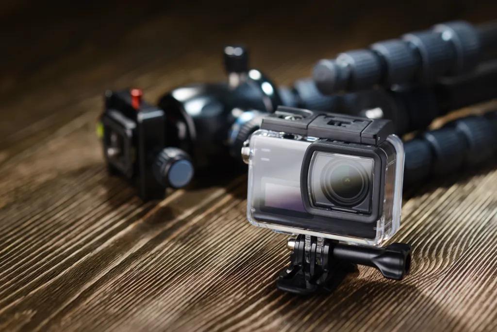 A black action camera in a clear waterproof case and a black tripod on a wooden surface.