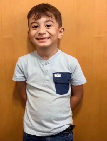 A smiling young boy in a light blue shirt with a dark blue pocket.