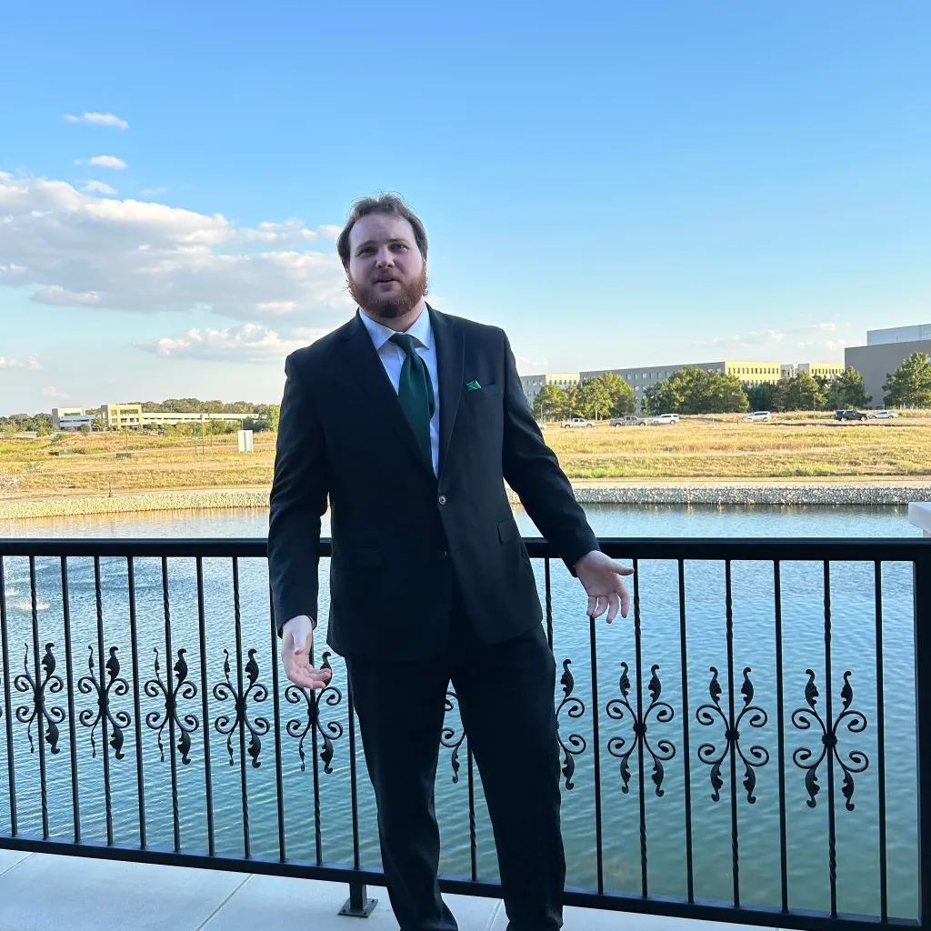 Braden Uhlmann, a man with a beard, wearing a suit and green tie, stands on a balcony overlooking a body of water with buildings in the background.