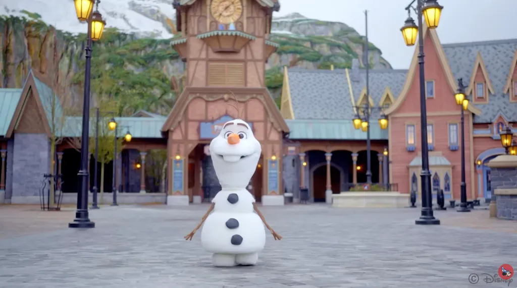 The character Olaf from Frozen standing in a town square in front of buildings and mountains.