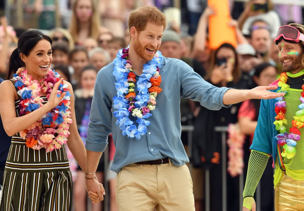 Meghan Markle, in a brown and white striped dress, and Prince Harry, in a blue shirt and khaki pants, wearing leis and holding hands while Harry gestures to a man in a neon outfit.