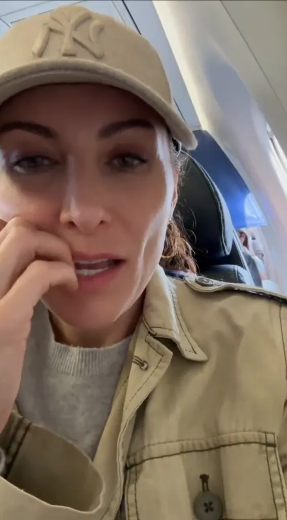 A woman in a New York Yankees baseball cap and trench coat on an airplane, looking distressed.