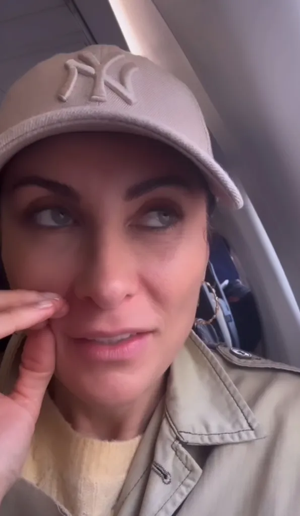 Laura Benanti in a beige baseball cap with the New York Yankees logo, wearing a beige trench coat over a yellow sweater, touching her face with her fingers, and looking away.