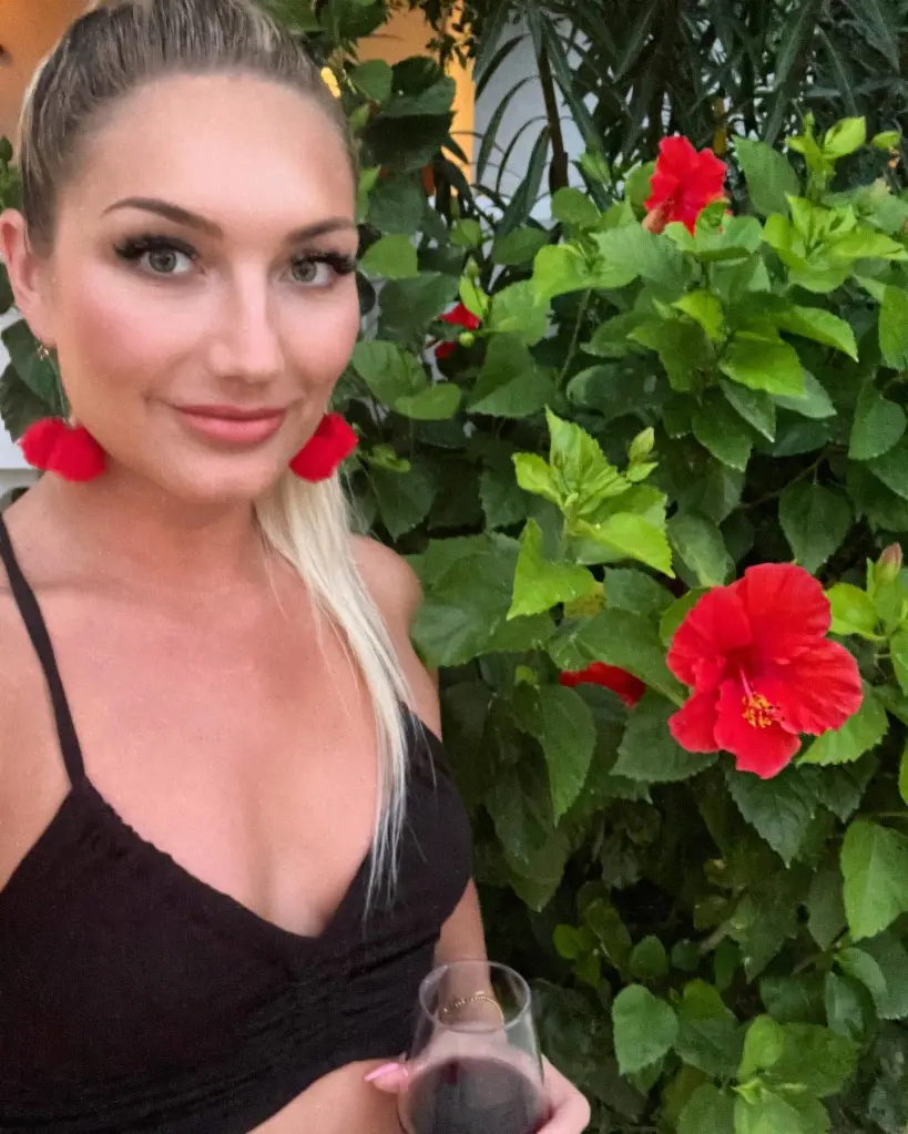 Brooke Hogan posing in front of a hibiscus bush while holding a glass of wine.