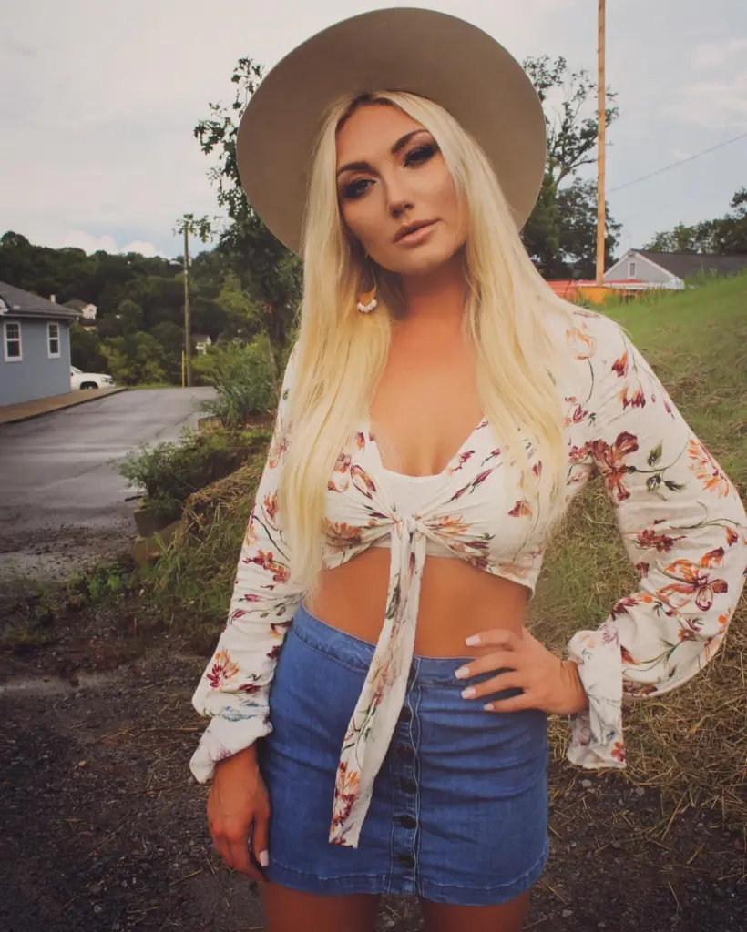 Brooke Hogan posing in a white floral top, denim skirt, and tan hat.