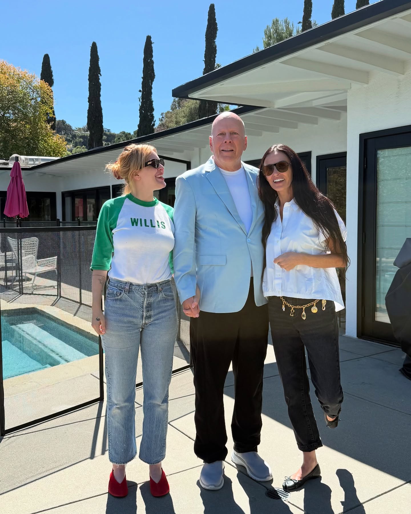 Bruce Willis, Demi Moore, and Rumer Willis stand side-by-side, holding hands and smiling for a photo.
