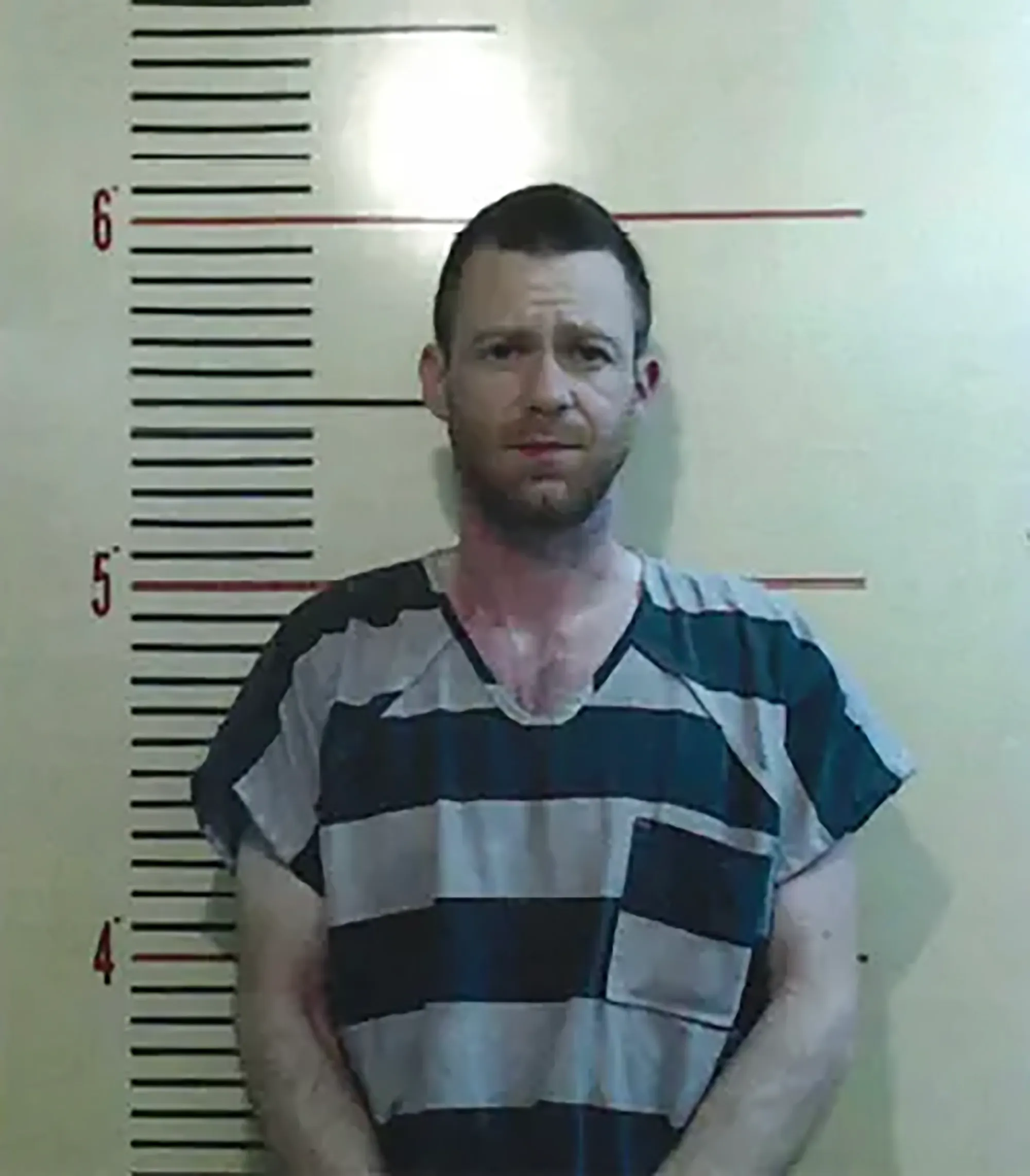Mugshot of Bug Hall, a man with short brown hair and a beard, wearing a striped prison uniform, standing against a height chart.