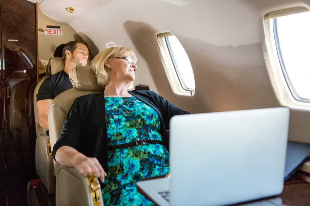 Two business travelers on a private plane: a woman working on a laptop, and a man sleeping in the seat behind her.
