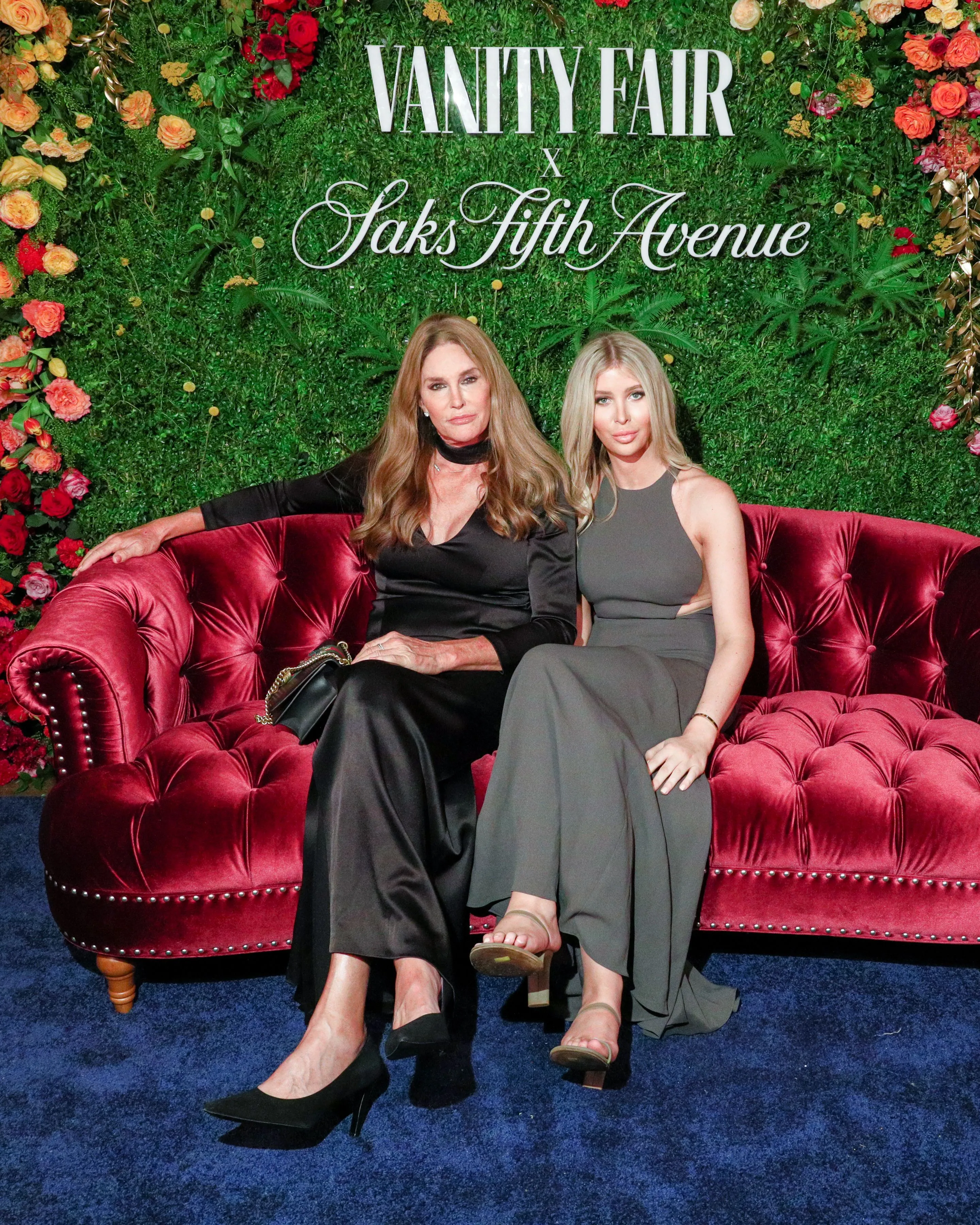 Caitlyn Jenner and Sophia Hutchins sitting on a red velvet couch in front of a green floral backdrop at an event.