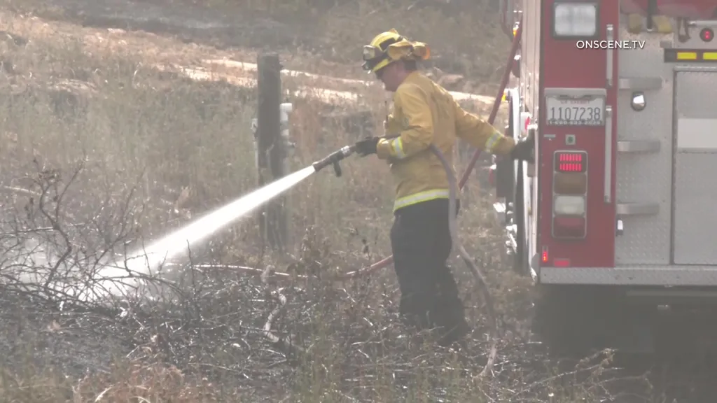 A firefighter spraying water onto smoldering brush next to a fire truck.