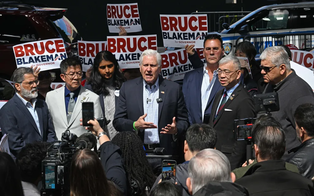 Bruce Blakeman speaks at a protest in Bath Beach, Brooklyn, surrounded by supporters holding signs for his gubernatorial campaign.