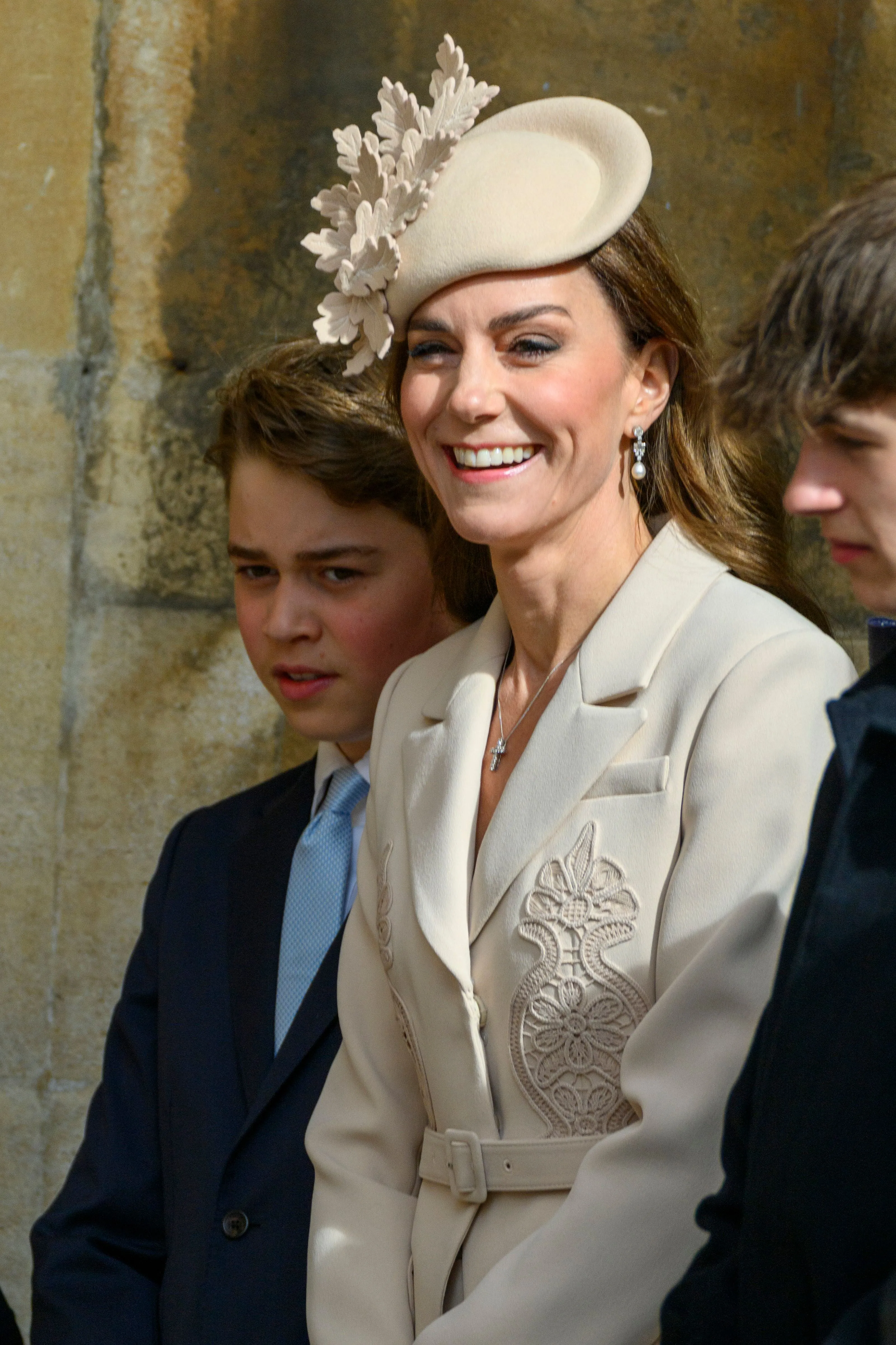Catherine, Princess of Wales, smiles wearing a cream-colored coat dress with a matching hat adorned with leafy details, accompanied by her son in a suit and light blue tie.