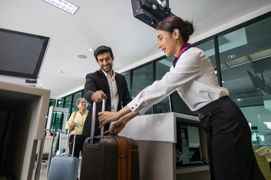 A businessman checking in a large bag with an airline ground crew member at a departure gate.