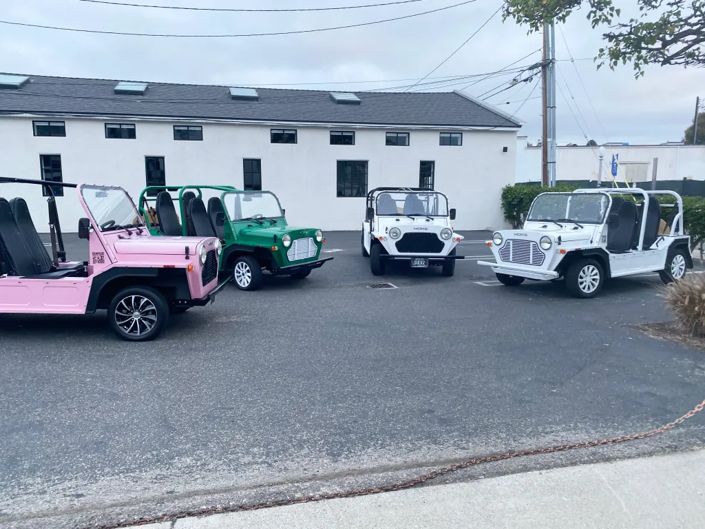 Four Moke electric vehicles, one pink, one green, and two white, parked outside.