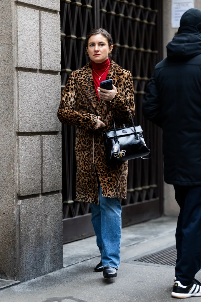 Chloe Malle wearing a leopard print coat, red turtleneck, blue jeans, and black boots, holding a black handbag and phone, at Milan Fashion Week.