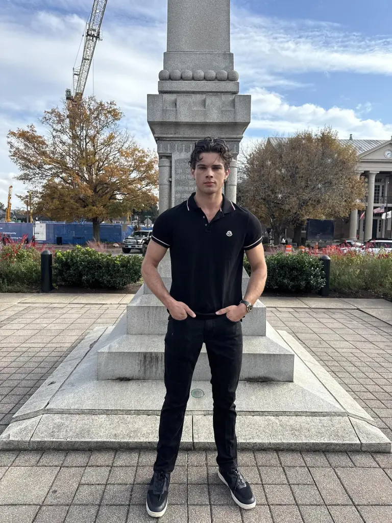 Braden Peters, a male model, poses in a black polo shirt and black jeans in front of a stone monument.
