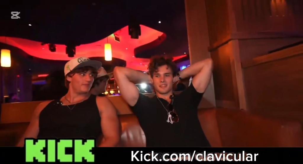 Two men at a club, one wearing a tank top and a baseball cap, the other wearing a t-shirt and sunglasses hanging from his neck.