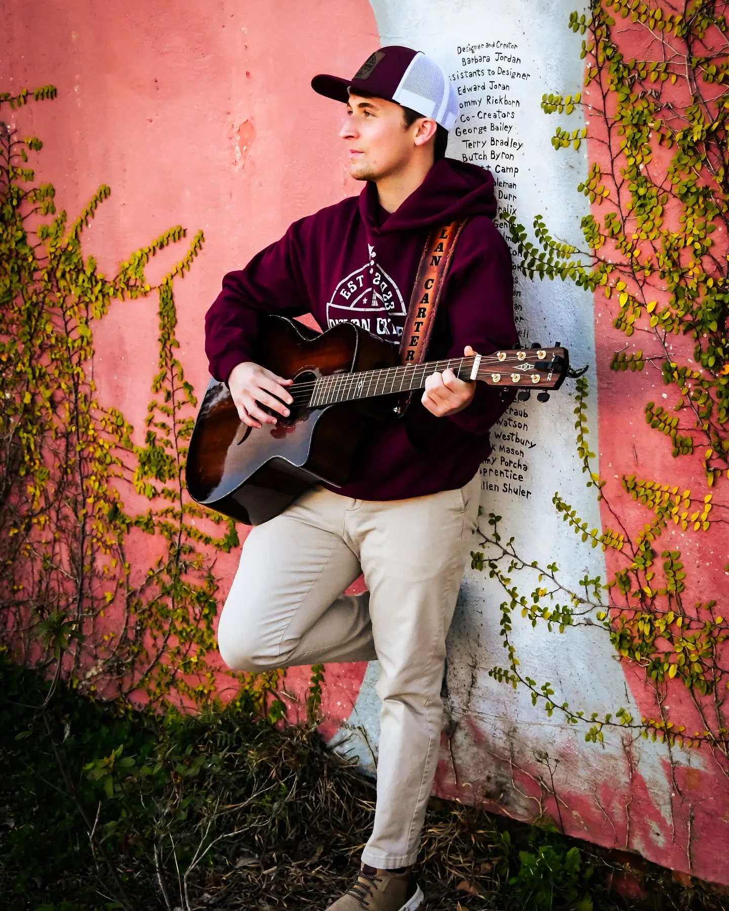 A man in a burgundy hoodie and hat holds a guitar and leans against a pink and white wall covered in vines.