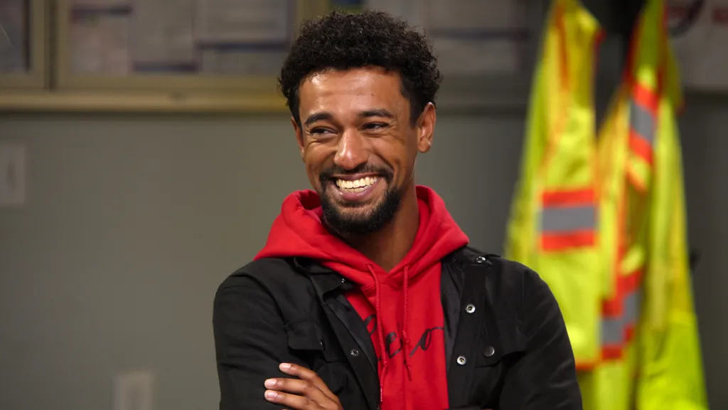 A smiling young man in a red hoodie and black jacket.