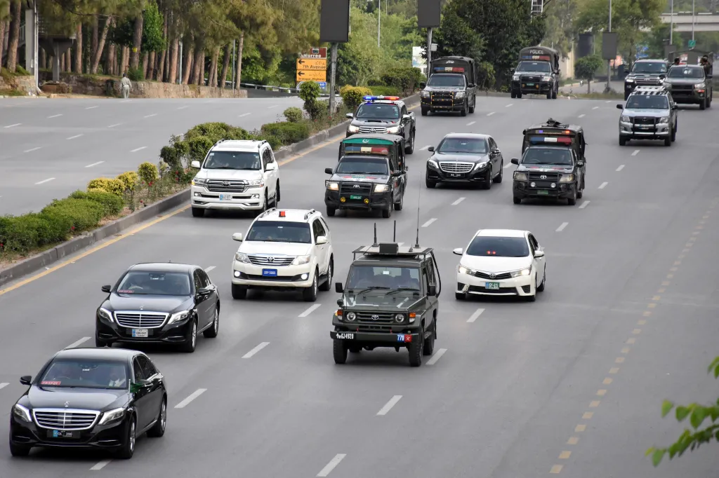 A convoy of black sedans, SUVs, and police trucks driving on a multi-lane road in Islamabad, Pakistan.