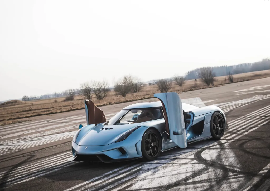A light blue Koenigsegg Regera with both doors open, parked on an asphalt track with tire marks.