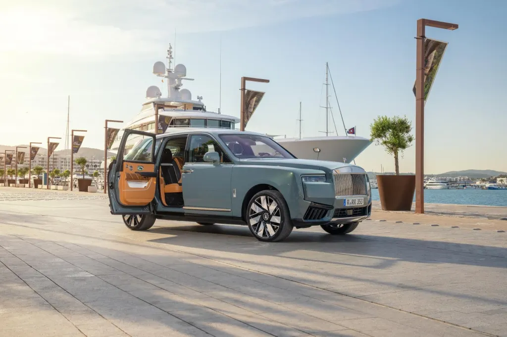 A Rolls-Royce Cullinan with an open door at a pier with yachts in the background.