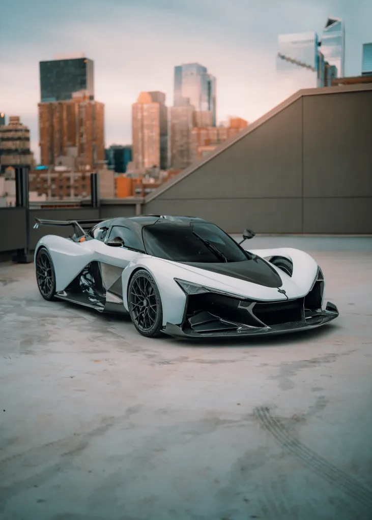 The Zenvo Aurora supercar, in white and black with exposed carbon fiber, is parked on a concrete rooftop with a city skyline in the background.