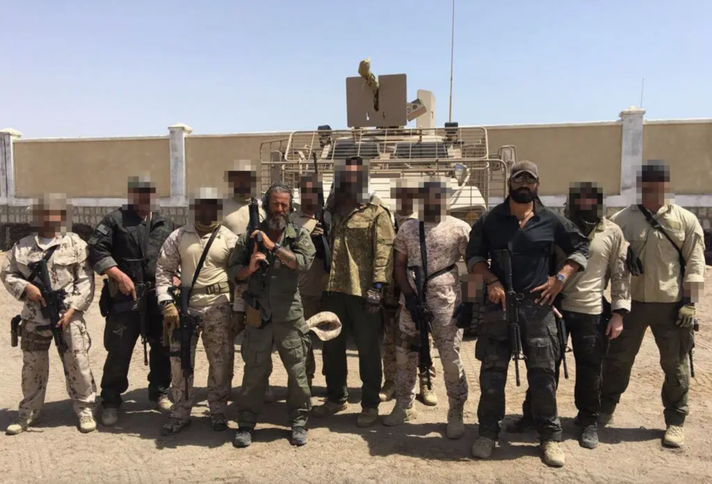 A group of armed men, some with blurred faces, posing in front of an armored vehicle.