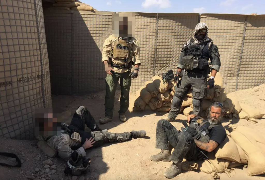 Four mercenaries in military gear, two standing and two sitting or lying down, in a sandbag-reinforced enclosure.