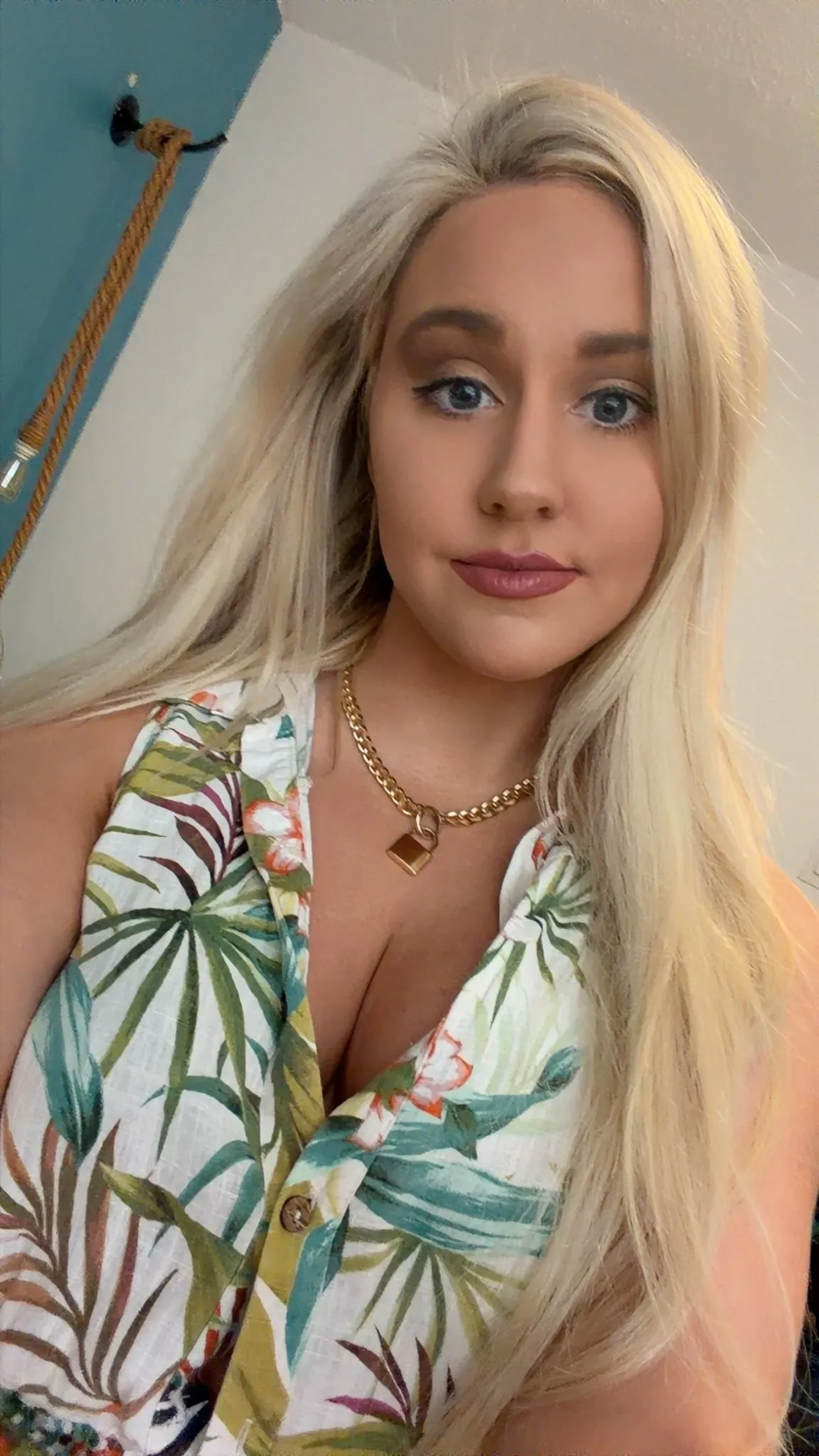 Crissy Froyd woman with long blonde hair, light blue eyes, and pink lipstick looks directly at the camera. She wears a floral print top with green leaves and a gold chain necklace with a padlock pendant.