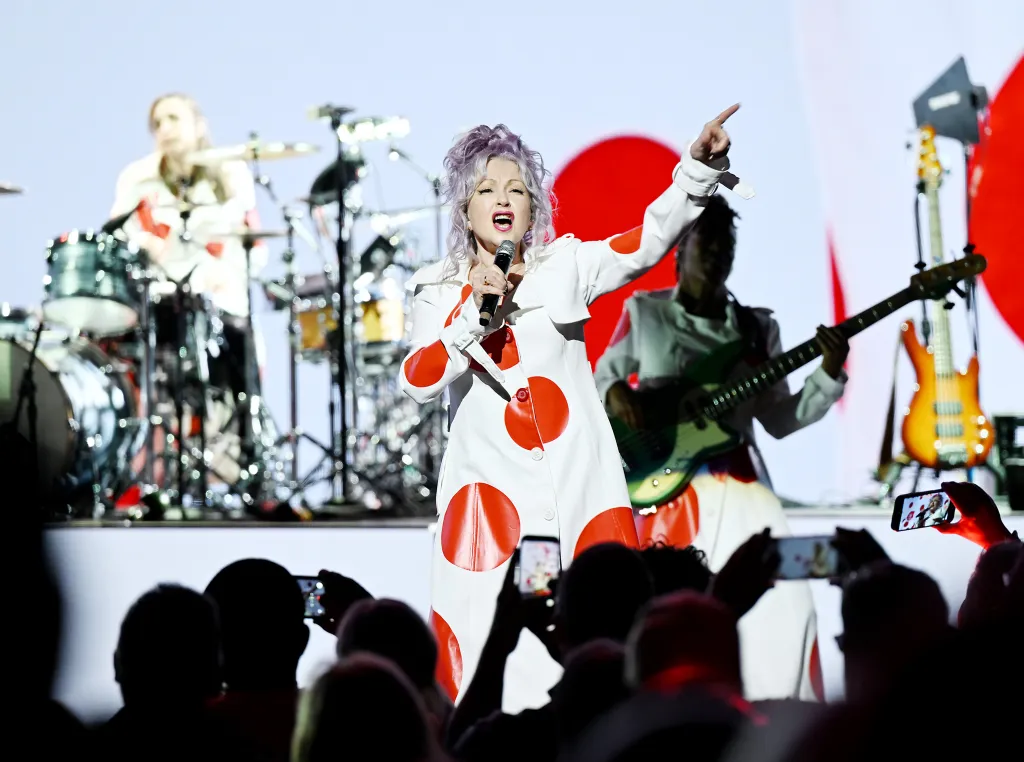 Cyndi Lauper performing live in a white coat with large red polka dots, singing into a microphone, with her band members and audience visible.